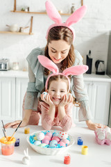 happy mother looking at bunny ears on daughter near easter eggs