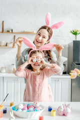 Happy mother touching bunny ears on daughter covering eyes with chicken eggs near tulips