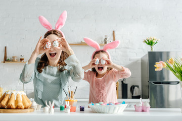 excited mother and daughter in bunny ears covering eyes with painted chicken eggs near tulips and easter bread