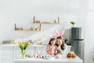 happy kid and mother with bunny ears near chicken eggs, decorative rabbits, easter bread and tulips