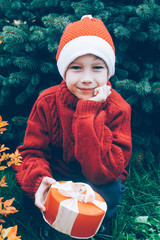 A boy in a red Santa suit holding a gift on the background of the Christmas tree. Sad Santa Claus.