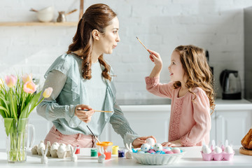 side view of cheerful mother and happy daughter looking at each other near painted easter eggs and flowers