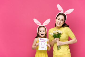 Cheerful mother in bunny ears holding greeting card with happy easter lettering near kid with tulips isolated on pink