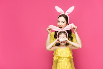 excited mother touching bunny ears of daughter covering eyes with easter eggs isolated on pink
