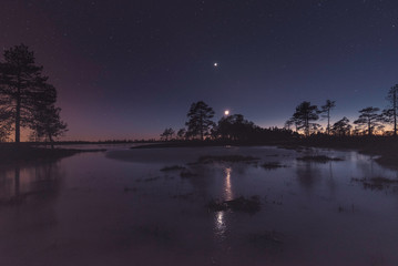 Fototapeta na wymiar Moon and Venus reflecting on frozen water bog with trees on the horizon