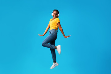 Joyful Black Woman In Headphones Jumping On Blue Studio Background