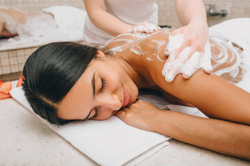 Obraz na płótnie Canvas Beautiful mixed race woman enjoys foam massage in a Turkish bath. Hammam worker washes woman skin