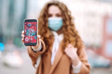 Girl in protective sterile medical mask on her face holds out the phone in quarantine city. Coronavirus cells on phone display. The concept of preventing the spread of the epidemic. 