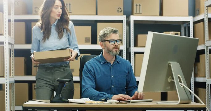 Caucasian post female worker bringing box to man who working at computer in delievery office. Male registering parcel and scanning bar code with scanner. Mailing office full of cardboard parcels.