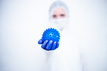 A virologist in a white mask and blue rubber gloves in a viral protective suit on a white background holds a blue ball in the form of .coronavirus, covid19,