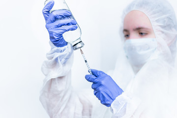 A virologist in a virus- proof suit and medical mask fills a syringe with pink medicine. A female nurse with a syringe in her hand on a white background.