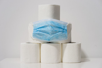 Front view on white toilet paper rolls stacked on the table in front of the wall background with blue protective mask during the virus epidemic situation concept