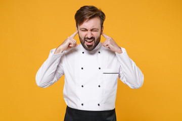 Irritated young bearded male chef cook or baker man in white uniform shirt isolated on yellow background. Cooking food concept. Mock up copy space. Keeping eyes closed, covering ears with fingers.