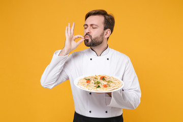 Young bearded male chef cook or baker man in white uniform shirt isolated on yellow background. Cooking food concept. Mock up copy space. Hold italian pizza on plate making okay taste delight sign.