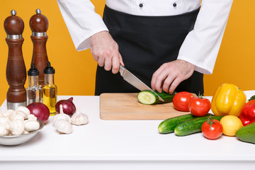 Cropped image of young male chef cook or baker man in white uniform shirt isolated on yellow background. Cooking food concept. Mock up copy space. Cooking salad at table, cutting cucumber on board.