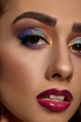 Naklejka premium Brunette woman with luxury makeup and perfect skin. Multi-colored eyeshadow, long eyelashes, glossy burgundy lips and white teeth. Close up