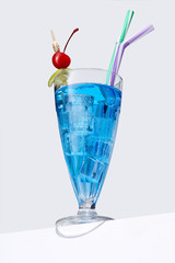 Blue Lagoon cocktail with ice cubes is contained in a milkshake glass with two straws, a lime slice and a cherry on the stick. The showy illustrative picture is made on the gray backdrop.
