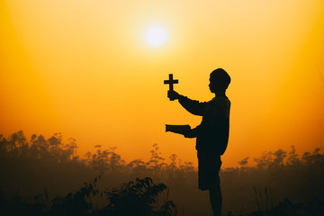 Young man holding bible ang christian cross on the mountain at sunset background. christian silhouette concept.