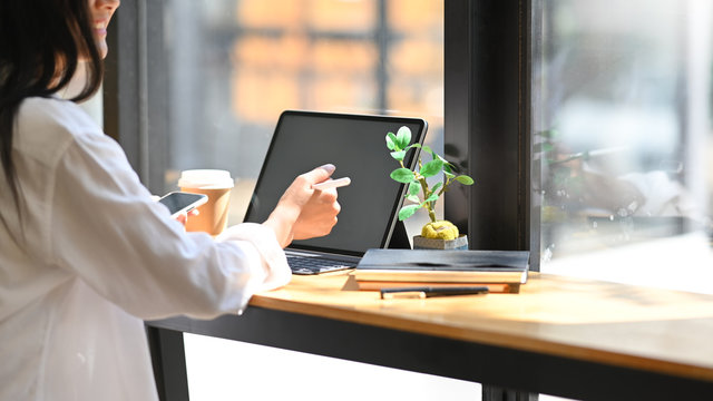 Cropped image of secretary woman's hand holding a smartphone in her hand while sitting and using a white blank screen computer tablet with keyboard case at the wooden working table in modern office.