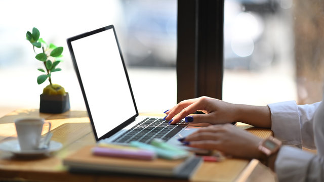 Cropped image of confident woman working with white blank screen computer laptop while sitting at the wooden working desk over modern office glass wall as background.
