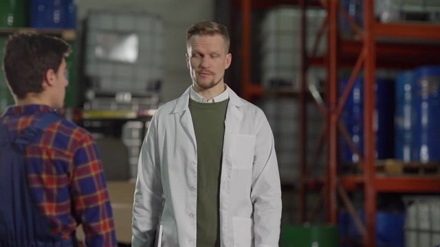 Slow motion medium shot of displeased inspector or manager in white coat asking questions to young material handler in industrial storage