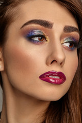 Brunette woman with luxury makeup and perfect skin is looking aside. Multi-colored eyeshadow, false eyelashes, glossy burgundy lips. Close up