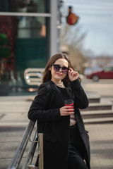 Obraz premium cheerful woman on a city street drinks coffee in a sunny day in sunglasses and looks away. coffee to go. Portrait of a girl with a drink in hands, vertical photo