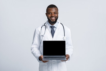 Black cheerful doc in white robe holding blank laptop screen