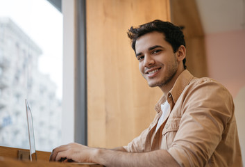 Successful programmer developer using laptop computer, sitting in modern office, looking at camera, smiling. Portrait of happy Indian freelancer working from home 