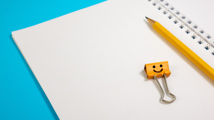 School Notepad with Yellow Pencil and Orange Smile Binder Clip. Office supplies on Blue Background. Open spiral notebook on table. Education concept. Back to school