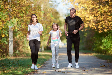 Mother, father and daughter wearing sportswear are jogging in autumn park alley, active family, warm and sunny weather