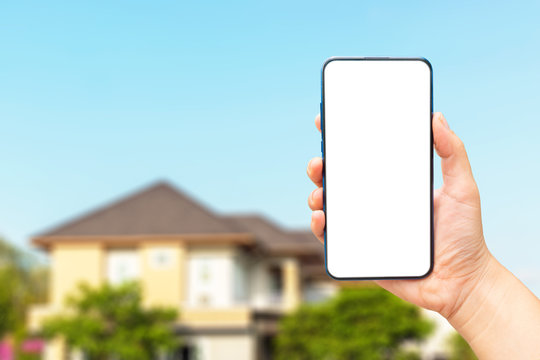 Mock up image of A hand holding a blank screen of smartphone on modern house blurred​ background.