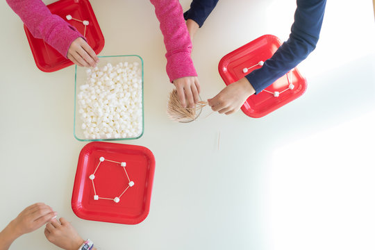 Directly above view of young children making geometric shapes with marshmallows and toothpicks as part of STEM lesson