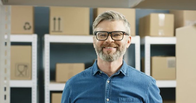 Close up of Caucasian handsome postman in glasses taking carton box with bar code from shelf and smiling in post office store. Portrait of man postal worker with happy smile and parcel in hands.