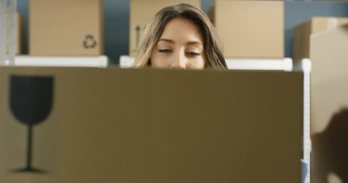 Close up of Caucasian beautiful woman taking carton box with bar code from shelf and smiling in post office store. Portrait of pretty female postal worker with happy smile and parcel in hands.