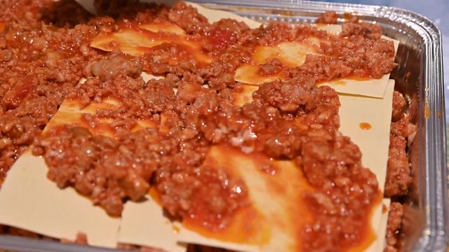 Homemade preparation of lasagna. Close-up image of the full pan: the hand, with a spoon, spreads the meat sauce on the pasta sheets to complete the preparation for the subsequent cooking.