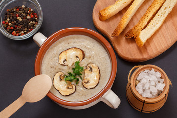 Cream soup with mushrooms in a porcelain bowl, spices and toasts on a black background.