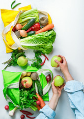 Textile shopping bags full of colorful vegetables and fruit on white background, woman's hands unpacking bags