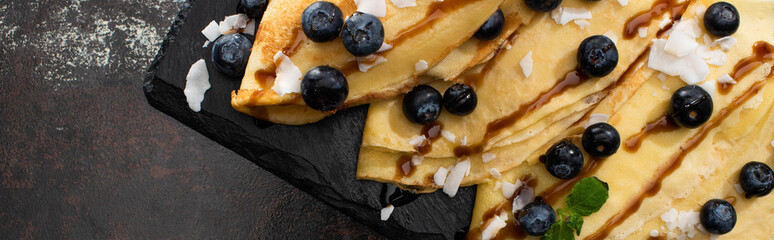 top view of tasty crepes with blueberries, mint and coconut flakes served on board on textured background, panoramic shot