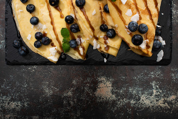 top view of tasty crepes with blueberries, mint and coconut flakes served on board on textured background