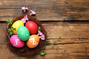 Colorful Easter eggs in decorative nest with flowers on wooden background, flat lay. Space for text