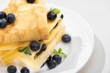 close up view of tasty crepes with mint and blueberries on plate on white background