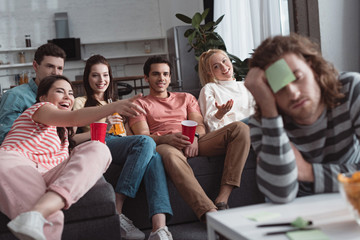 cheerful girl pointing with hand at bored man with sticky note on forehead while playing name game with friends