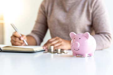 Young business women are saving money in a piggy bank and recording financial income-expenditures to prepare investments in real estate, Tax deduction and cost calculation concept.