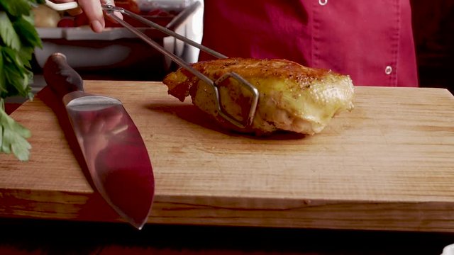 Chef putting a cooked chicken breast on a wooden cutting board with metal tongs