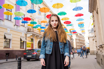 Beautiful girl walking under umbrellas in the city. Cute teenage girl in black dress posing against a background of multicolored umbrellas alley. Spring sunny concept. 8 March. Happy Women's Day.