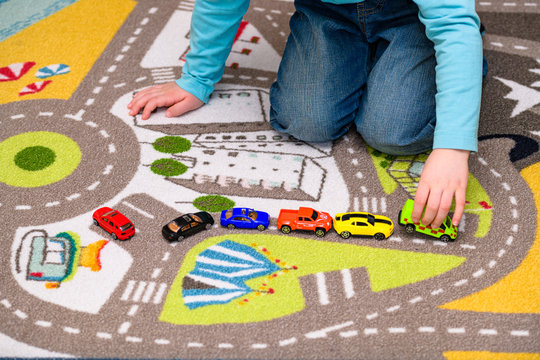 Five year old boy playing and lining up toy cars on a playing mat with roads. The cars have vivid colors and the boy is dressed in blue jeans and a blue shirt. He is holding a toy ambulance.
