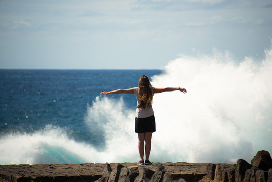 Giovane ragazza braccia aperte difronte all'oceano con gli schizzi e la spuma delle onde felice di viaggiare in libert&agrave; turista a Tenerife, Spagna