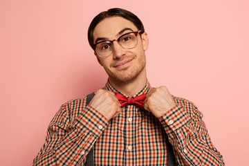 smiling male nerd in eyeglasses adjusting bow tie on pink