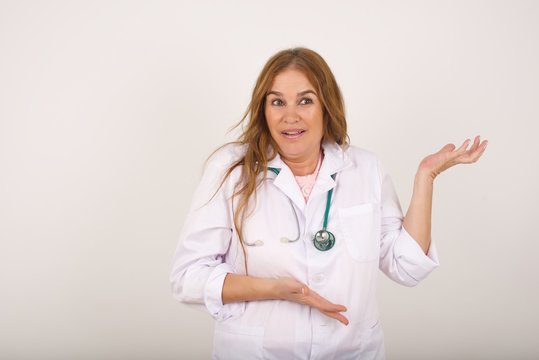 Beautiful European doctor girl pointing aside with both hands showing something strange and saying: I don't know what is this. Standing against gray background. Advertisement concept.